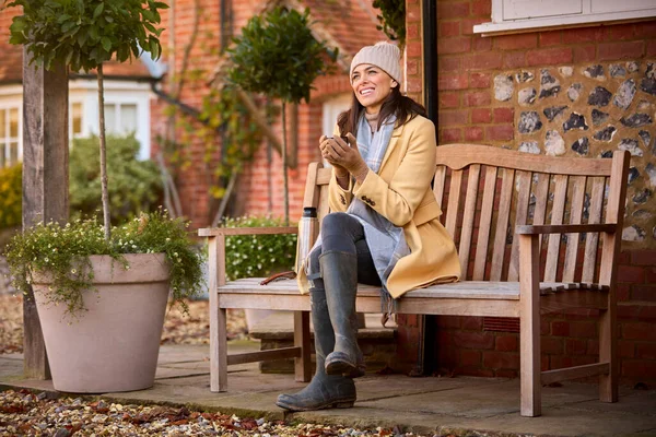 秋に家の外のベンチに座ってフラスコから熱い飲み物で帽子とスカーフを身に着けている女性 — ストック写真