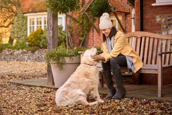 秋の散歩に準備ができてペット犬と家の外のベンチに座って帽子とスカーフを身に着けている女性 — ストック写真