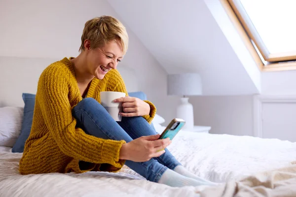Młoda Kobieta Siedzi Łóżku Domu Śmiejąc Się Patrząc Telefon Komórkowy — Zdjęcie stockowe