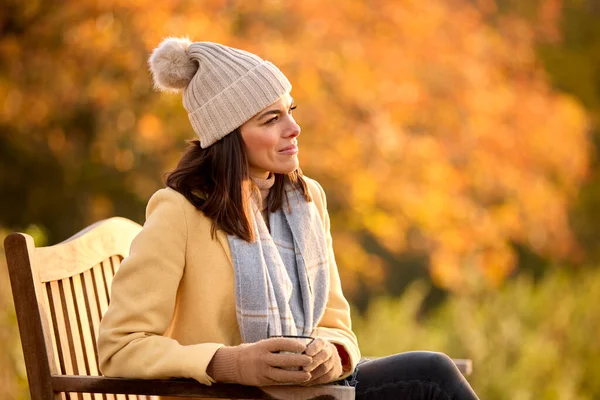 秋の田園地帯を歩く上でベンチに座って熱い飲み物と帽子とスカーフを身に着けている女性 — ストック写真