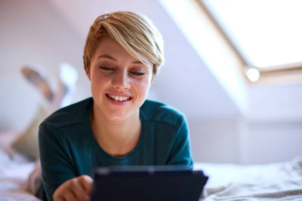 若い笑顔の女性が自宅でパジャマを着てベッドの上で寝そべっているデジタルタブレットを見て — ストック写真