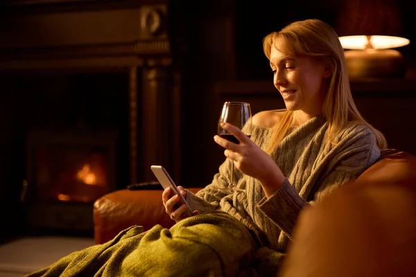 坐在沙发上的女人 炉火熊熊的客厅里 带着空白屏风的手机 手里拿着酒杯 — 图库照片