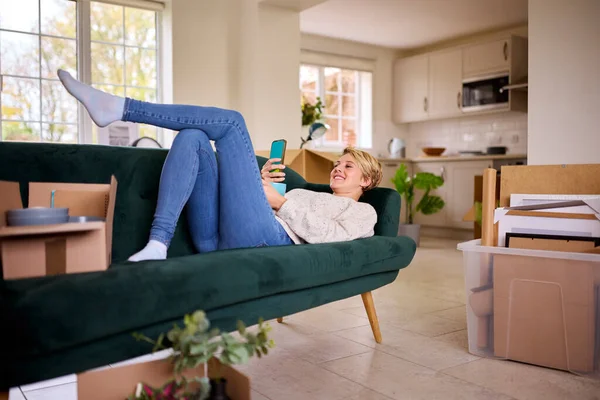 携帯電話を使ってソファでリラックスする女は 新しい家に引っ越してから箱を開けることで囲まれた — ストック写真