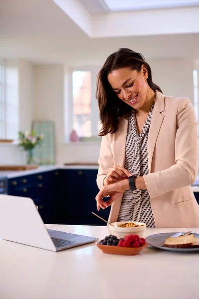 在家里的女商人 带着手提电脑在厨房柜台边吃早餐检查智能手表 — 图库照片