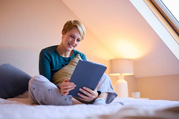 自宅でパジャマを着たままベッドに座っている若い笑顔の女性の夕景デジタルタブレットを見て — ストック写真