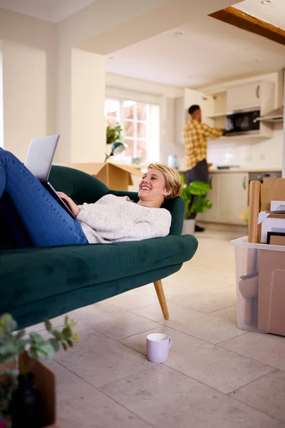 女性がラップトップを使用してソファに横たわっているように 新しい家に移動した後のカップルのアンパックボックス — ストック写真