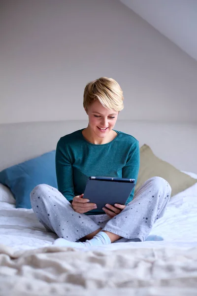 若い笑顔の女性は自宅でパジャマを着てベッドに座っているデジタルタブレットを見て — ストック写真