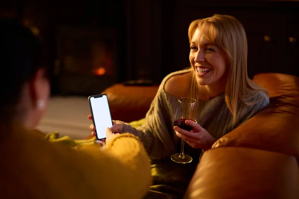 两个女性朋友在沙发上放松一下 一边看手机 一边喝着热腾腾的红酒 — 图库照片