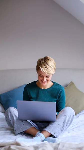 若い笑顔の女性は自宅でパジャマを着てベッドに座っているラップトップで作業 — ストック写真