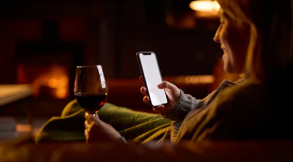 女人躺在沙发上 炉火熊熊的客厅里 用空白屏风手机拿着酒杯喝酒 — 图库照片