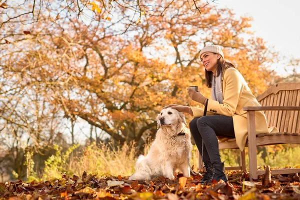 秋の田舎の散歩で犬とベンチに座って熱い飲み物と帽子とスカーフを身に着けている女性 — ストック写真