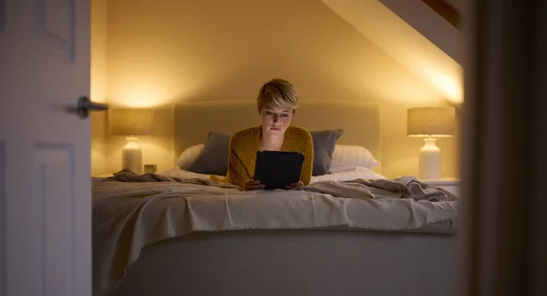自宅でベッドに寝そべっている若い女性の扉を通しての夕景デジタルタブレットを見て — ストック写真