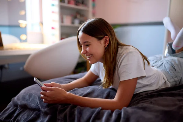 Χαμογελώντας Έφηβος Κορίτσι Ξαπλωμένη Στο Κρεβάτι Στο Σπίτι Κοιτάζοντας Κινητό — Φωτογραφία Αρχείου