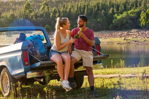 Couple With Backpacks In Pick Up Truck On Road Trip By Lake Drinking Beer