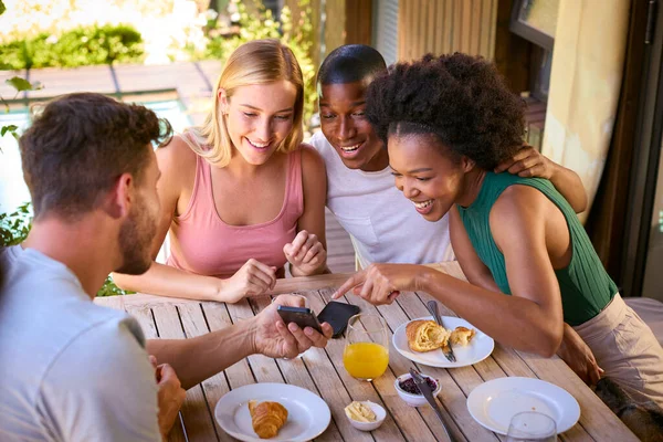 Group Of Smiling Multi-Cultural Friends Eating Breakfast Outdoors At Home Looking At Mobile Phone