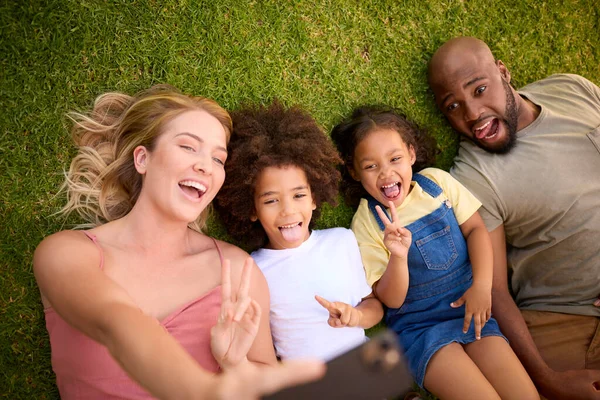 携帯電話でセルフィーを取る草の上に横たわる多人種家族のオーバーヘッドショット — ストック写真