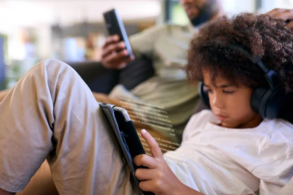 携帯電話および息子が付いている父親の近くでヘッドフォンが付いているデジタル タブレットの読書を使用して家 — ストック写真
