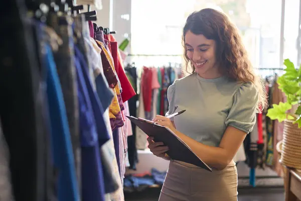 Female Owner Or Worker In Fashion Clothing Store Checking Stock With Clipboard