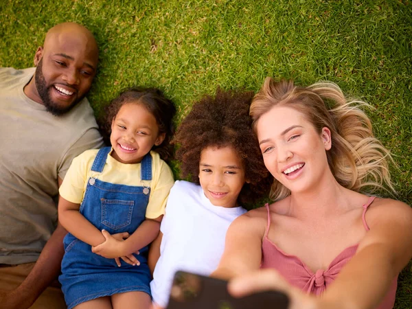 携帯電話でセルフィーを取る草の上に横たわる多人種家族のオーバーヘッドショット — ストック写真
