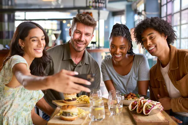 Group Of Friends Meeting Up In Restaurant Posing For Selfie On Mobile Phone With Food