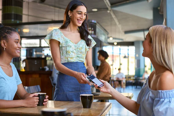 Woman In Coffee Shop Meeting Friend Paying Bill With Contactless Mobile Phone Payment