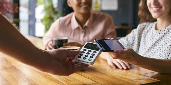 Woman In Coffee Shop Paying Bill With Contactless Mobile Phone Payment