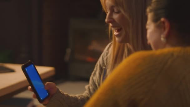 青い画面の携帯電話を見てソファーでリラックスする2人の女性の友人と居心地の良い火の前でワインを飲む — ストック動画