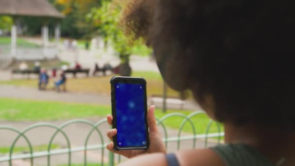 在城市公园里 穿着无线耳机 用蓝屏手机播放音乐或电影的女性从背后慢镜头观看的特写镜头 — 图库视频影像
