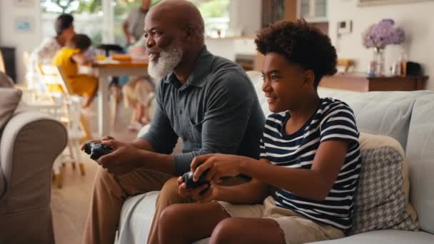 祖父と孫が家でソファーに座り コントローラーがバックグラウンドで複数の世代の家族と一緒にビデオゲームをプレイする スローモーションで撮影 — ストック動画