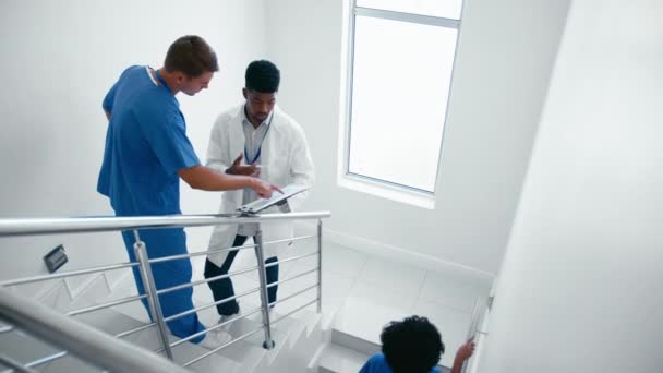 男医生身穿白色外套 与护士在医院楼梯上的洗澡间里讨论病人笔记 由女同事经过 动作缓慢 — 图库视频影像