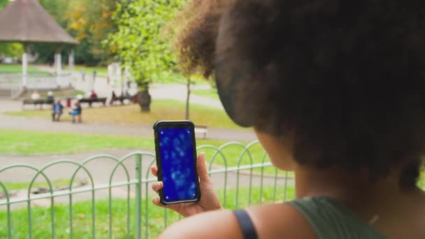 在城市公园里 穿着无线耳机 用蓝屏手机播放音乐或电影的女性从背后慢镜头观看的特写镜头 — 图库视频影像