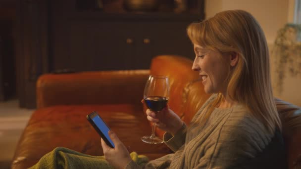 坐在沙发上 拿着毛毯的女人靠在沙发上 看着蓝色屏幕的手机 喝着一杯背景火热的葡萄酒 慢镜头拍摄 — 图库视频影像