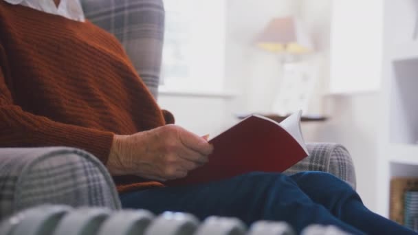 老妇人坐在扶手椅上 坐在家里的便携式散热器旁看书 动作缓慢 让人感到温暖 — 图库视频影像