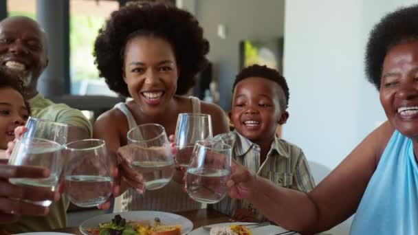 家庭でテーブルの周りに座っている多世代の家族は 食事でカメラに向かって水眼鏡で応援しています スローモーションで撮影 — ストック動画