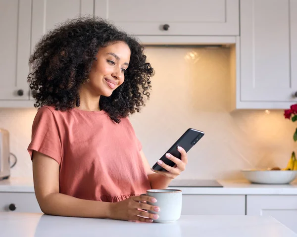 Young Woman Leaning On Kitchen Counter At Home Checking Social Media On Mobile Phone With Hot Drink