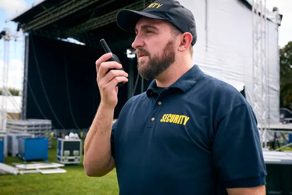 Security Team At Outdoor Stage For Music Festival Or Concert Talking Into Radio