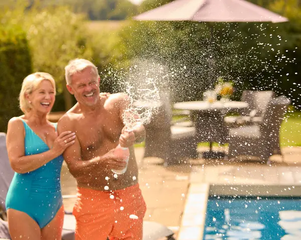 Senior Couple On Holiday In Swimming Costumes Opening Champagne By Hotel Swimming Pool
