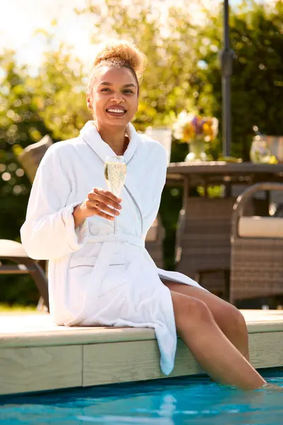 Woman In Robe Sitting Outdoors With Feet In Swimming Pool Drinking Glass Of Champagne At Spa