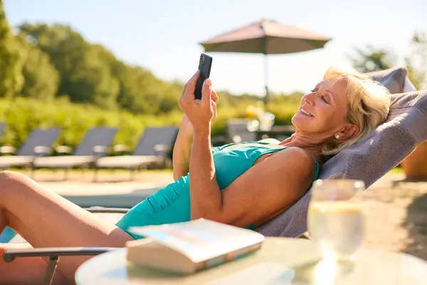 Senior Woman On Holiday In Swimming Costume Looking At Mobile Phone Relaxing By Hotel Swimming Pool