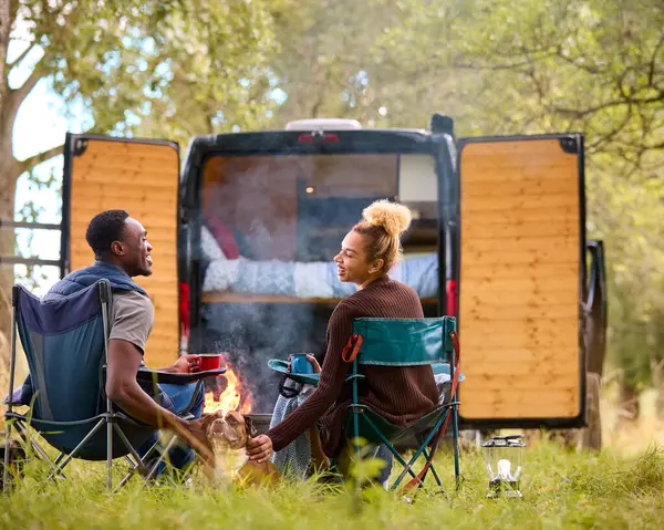 Rear View Of Couple Camping In Countryside With RV Drinking Coffee By Outdoor Fire With Pet Dog