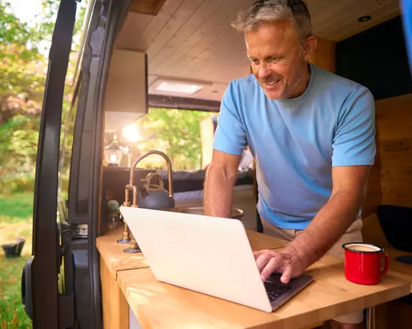 Senior Man On Camping Trip In Countryside Working Inside RV Using Laptop