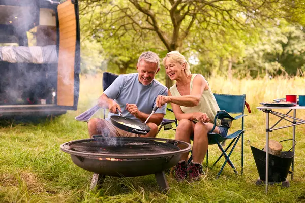 Senior Couple Camping In Countryside With RV Cooking Bacon And Eggs For Breakfast Outdoors On Fire