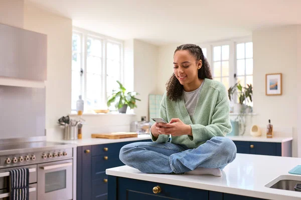 Teenage Girl At Home On Kitchen Island Connecting With Friends On Social Media Using Mobile Phone