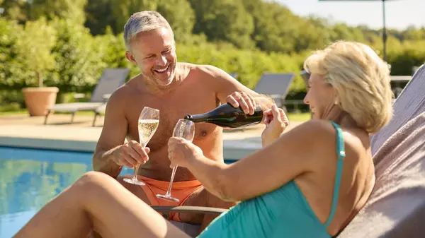 Senior Couple On Holiday In Swimming Costumes Pouring Champagne By Hotel Swimming Pool