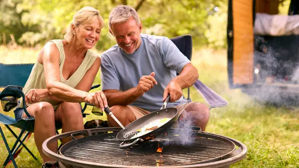 Senior Couple Camping In Countryside With RV Cooking Bacon And Eggs For Breakfast Outdoors On Fire