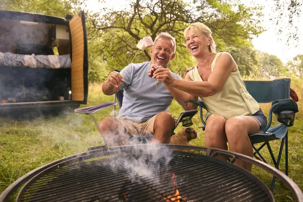 Senior Couple Camping In Countryside With RV Toasting Marshmallows Outdoors On Fire