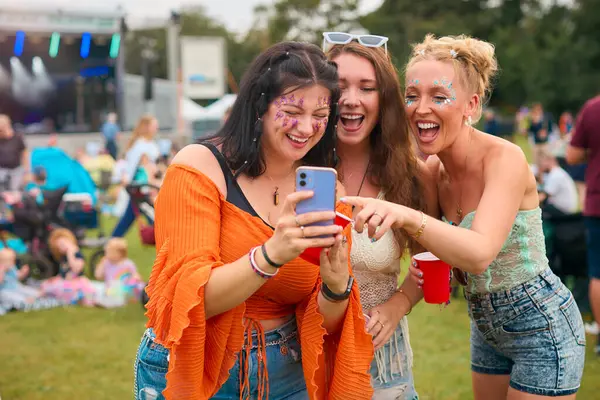 サマーミュージックフェスティバル ホールディングドリンクで携帯電話を見ているグリッターを着ている3人の女性の友人 — ストック写真
