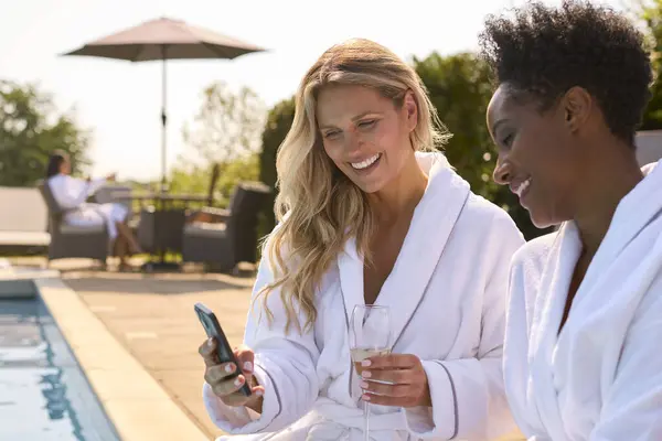 Two Mature Female Friends With Mobile Phone In Robes Outdoors By Pool Drinking Champagne On Spa Day
