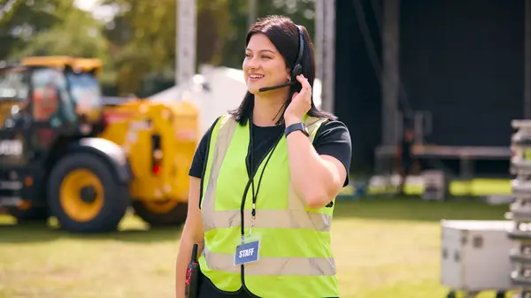 Female Production Worker Talking On Headset Setting Up Outdoor Stage For Music Festival Or Concert