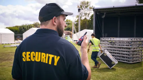 Rear View Of Security Team At Outdoor Stage For Music Festival Or Concert Talking Into Radio